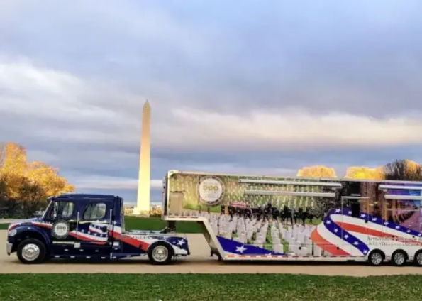 Blue bus with red, white, and blue American flag graphics connected to a large trailer showing a marching band, with the Washington Monument in the background.