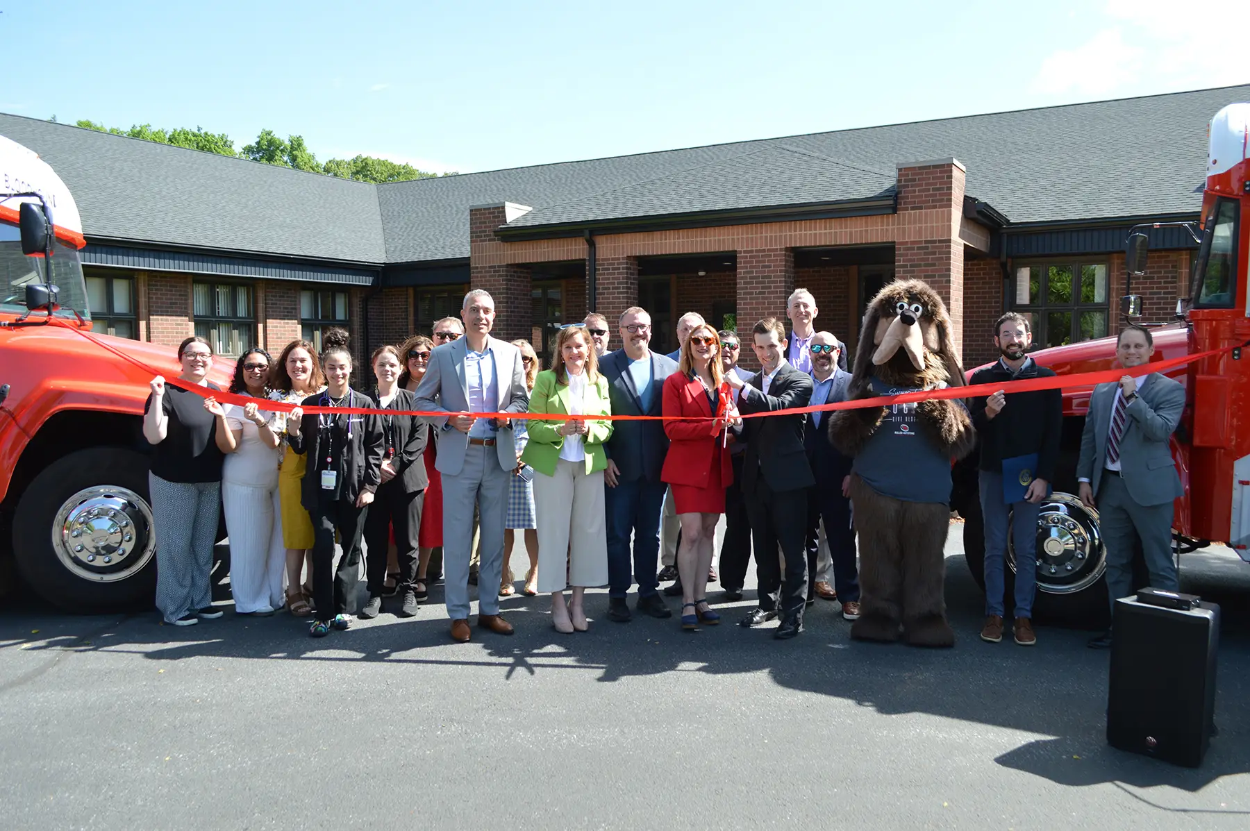 Group of professionals cutting a red ribbon at a grand-opening ceremony in front of a brick office building, with red trucks on either side and a costumed mascot nearby.
