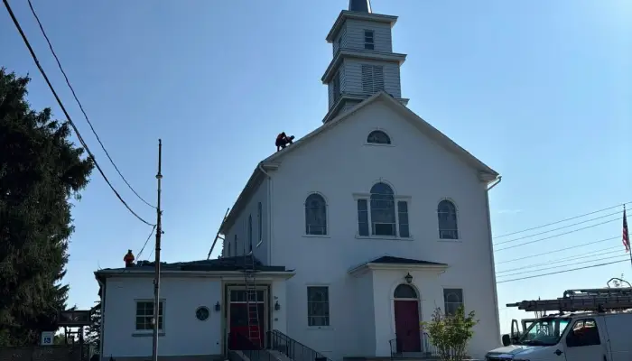 White church with red door and tall steeple, a worker on the roof on a sunny day.