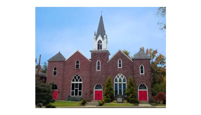 Front view of a brick church with arched windows, four red doors, and a tall white steeple against a blue sky.