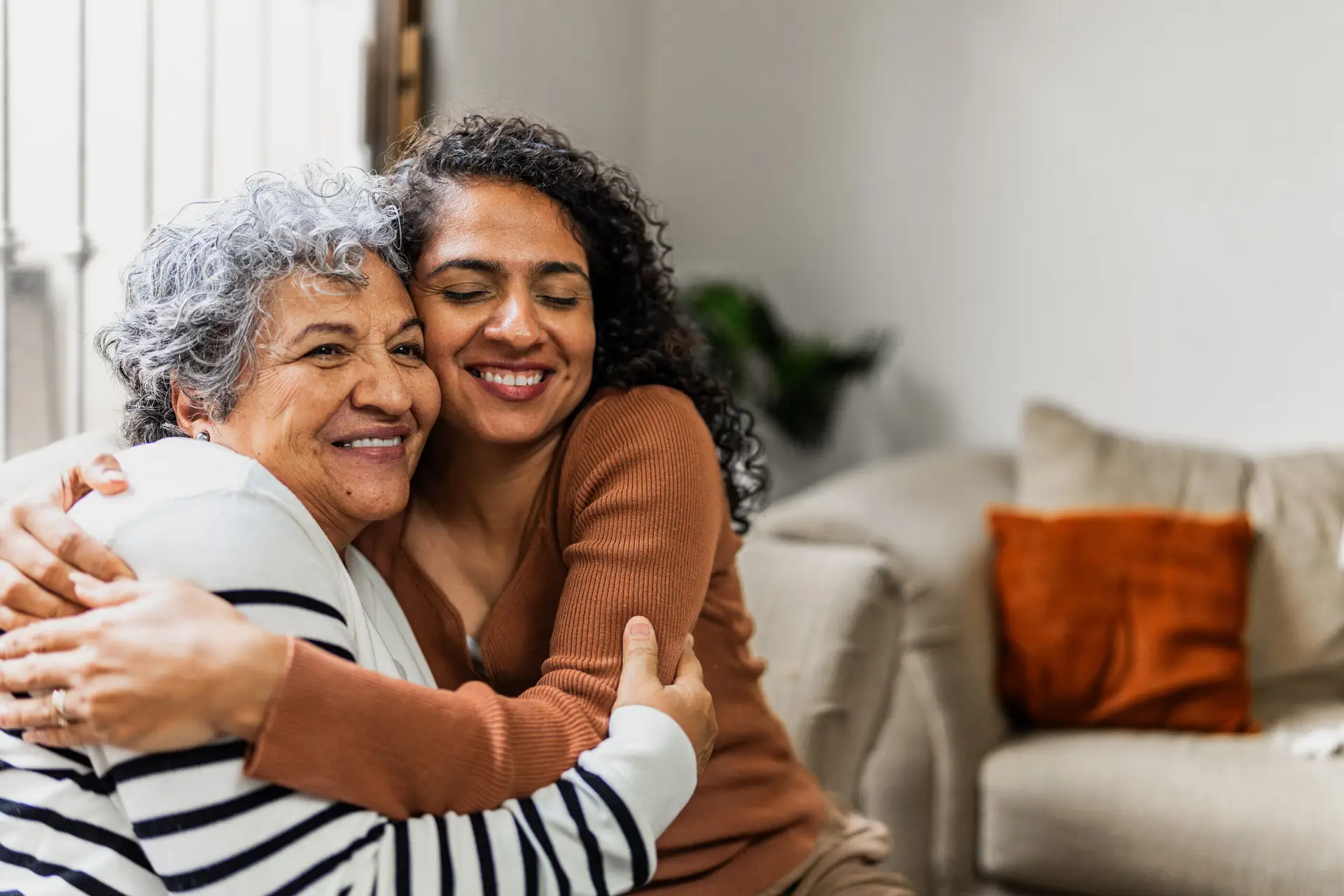 Two women hug warmly on a beige sofa in a bright living room, both smiling with their eyes closed.