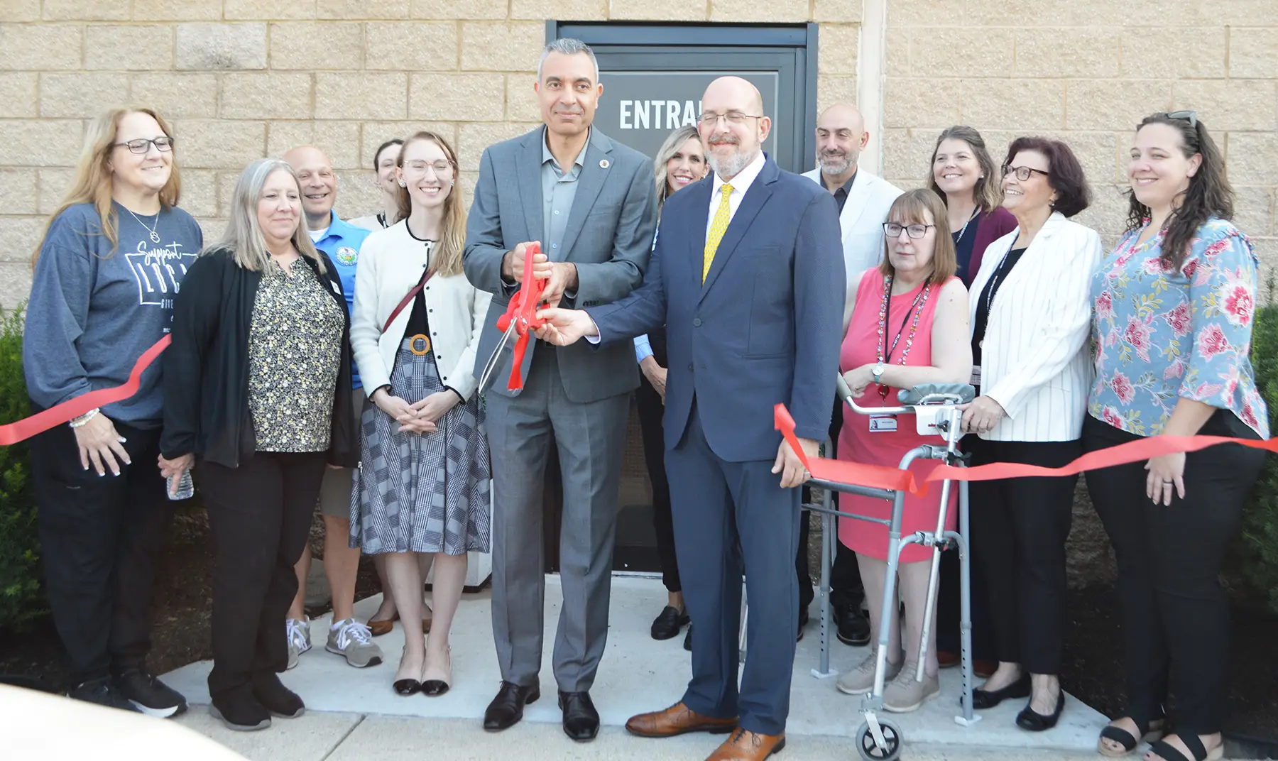 Group of adults posing for a ribbon-cutting ceremony outside a building entrance. Two men in suits cut the red ribbon while others smile and watch.