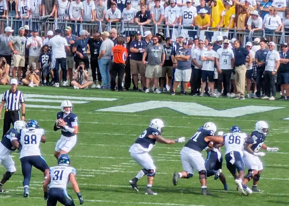 Football game in progress with white-and-navy players blocking near the line while fans watch from the stands.