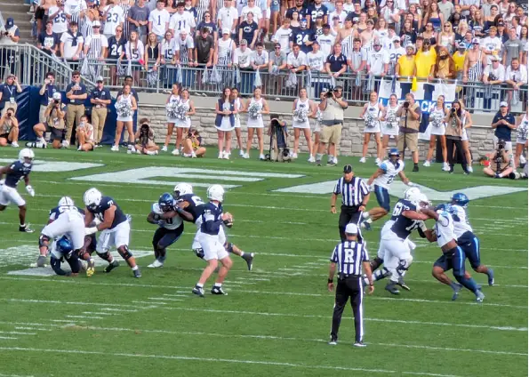 American football game in progress on a green field, players in dark and white uniforms clash near the line of scrimmage as referees watch; a large crowd of spectators fills the stands in the background.