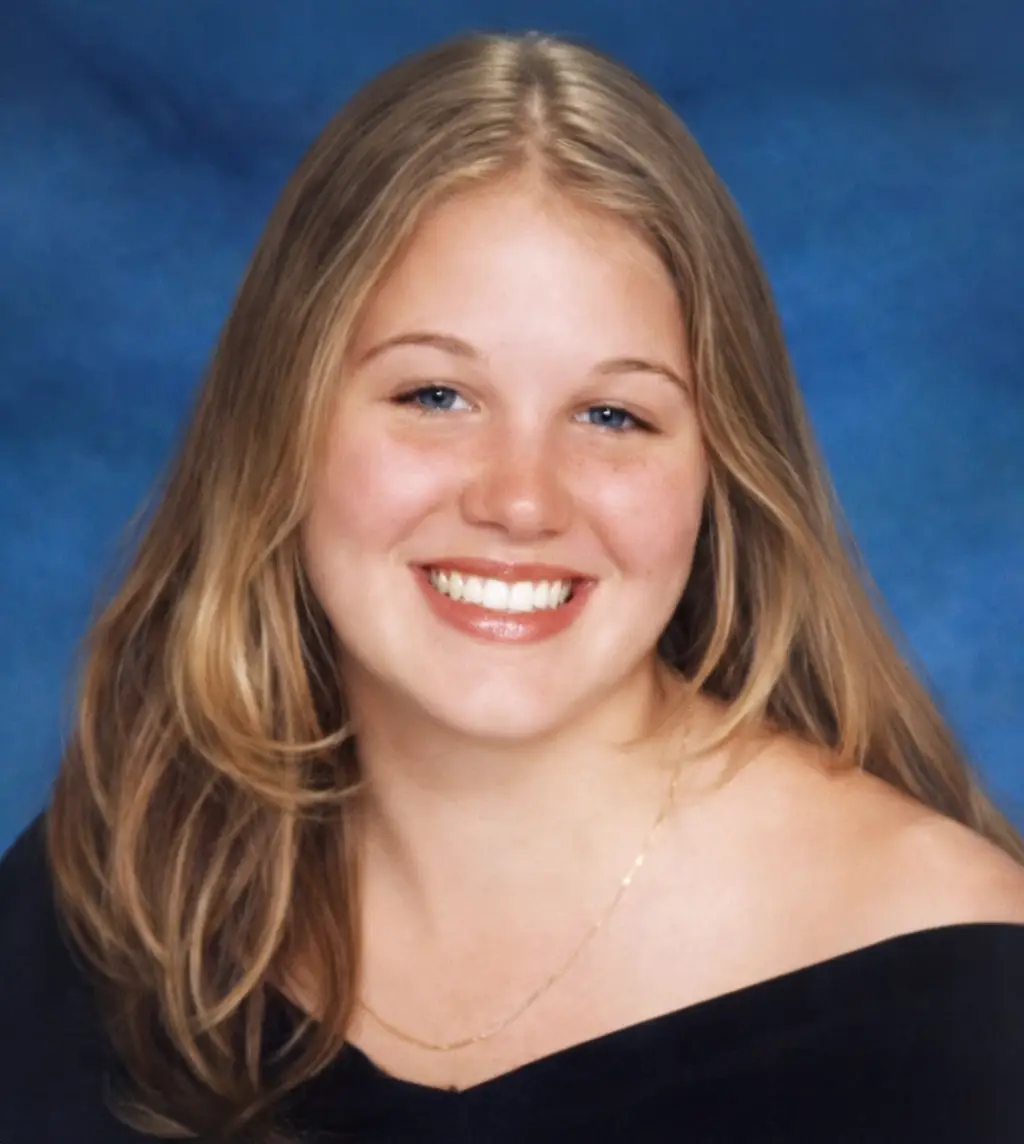 Smiling young woman with long blonde hair posing for a portrait against a blue backdrop, wearing an off-the-shoulder black top and a delicate gold necklace.