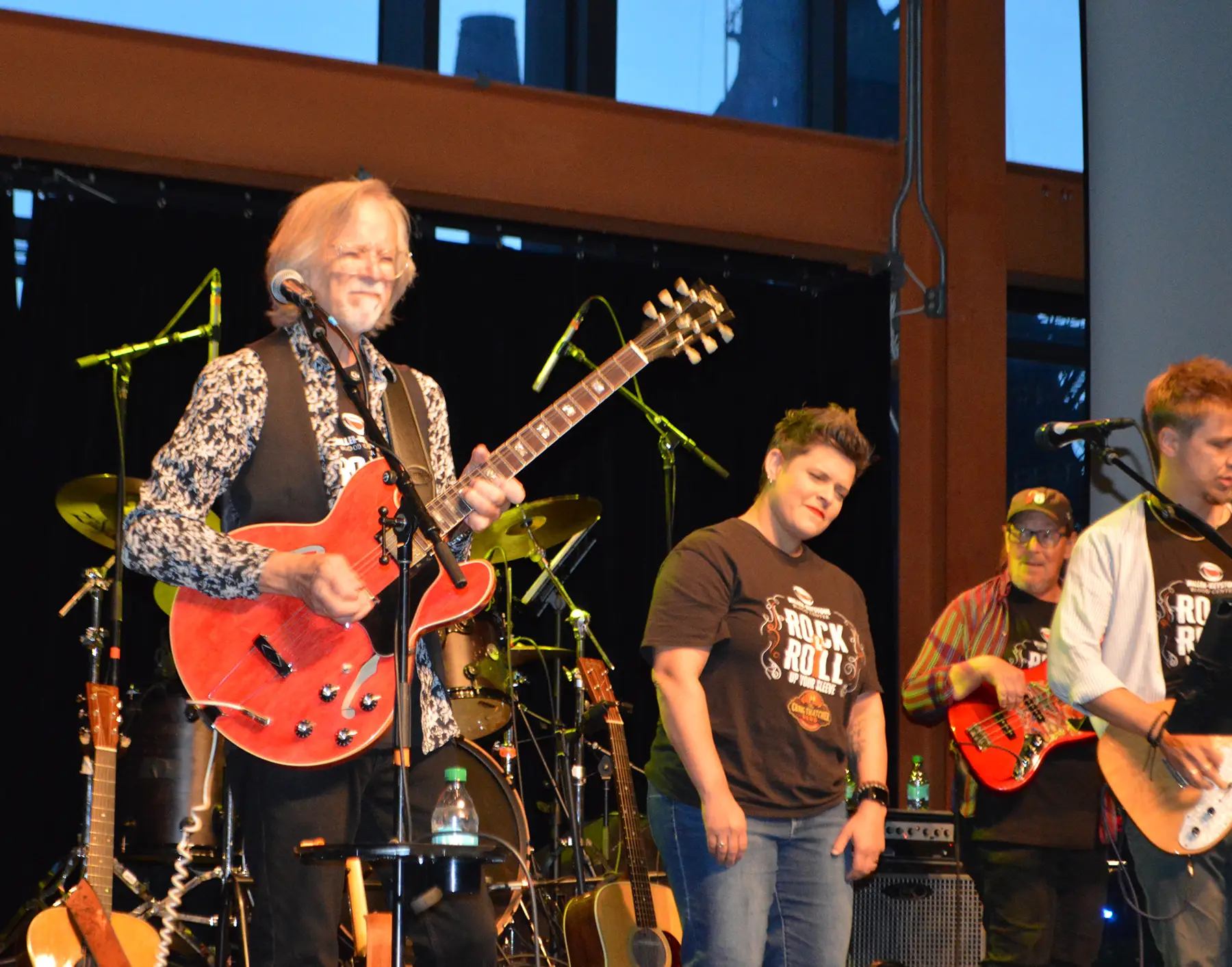 Guitarist with a red semi-hollow guitar on stage, wearing a patterned shirt and vest, smiling as he plays.